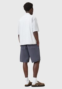 Camicia a maniche corte bianca e azzurro chiaro a righe, pantaloni corti grigi con tasche e zoccoli marroni con cinturini, indossati con calze bianche.