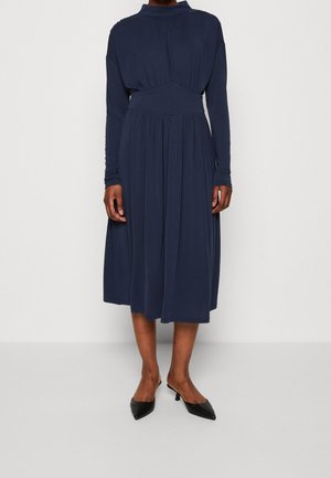 Person wearing a navy blue long-sleeve midi dress with high neckline and black low-heeled pointed-toe shoes against a plain background.