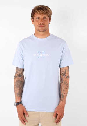 T-shirt en coton bleu clair avec un logo "Oxbow" blanc entouré de motifs de feuilles. Manches courtes et col rond, avec des tatouages visibles sur les bras.