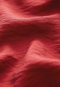 Červená látka s jemnou texturou a mírným leskem, vykazující jemné vlnění, což naznačuje lehký materiál vhodný pro oděvy nebo domácí dekoraci.