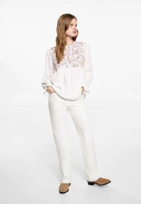 Bijela vezeno bluza s cvjetnim uzorcima i dugim rukavima, u kombinaciji s krem hlačama ravnog kroja i smešnim čizmama do gležnja. Glatke teksture tkanina.