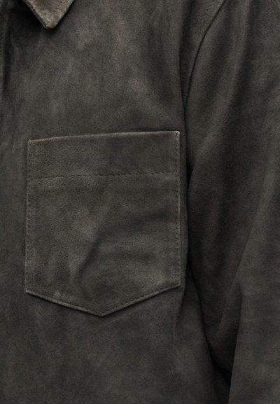 Cămașă gri închis din imitație de piele întoarsă, cu guler ascuțit și o buzunar la piept cu partea superioară dreaptă. Textura pare moale și netedă.