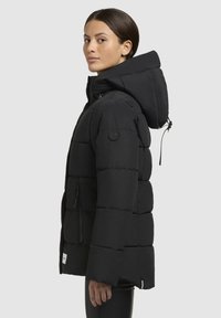 Veste en duvet noire avec capuche, affichant un design matelassé, des poches zippées et un patch logo sur la manche. Texture lisse et silhouette ajustée.