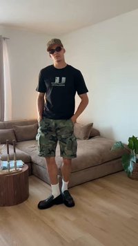 Un homme portant des lunettes de soleil, un t-shirt noir, un short camouflage, des chaussettes blanches et des chaussures noires se tient dans un salon avec un canapé marron et une plante.