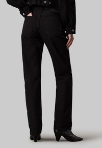 Pantalones de denim negro con corte recto, con dos bolsillos traseros, detalles de costura sutiles, y combinados con calzado negro puntiagudo.