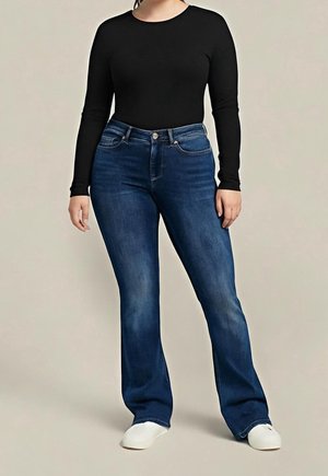 Vrouw draagt een zwarte top met lange mouwen, donkerblauwe recht gesneden spijkerbroek en witte sneakers, staand tegen een eenvoudige achtergrond.