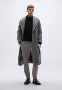 Szürke halszálkamintás hosszú kabát fekete kötött pulóverrel, világosszürke nadrággal és fekete bőrcipővel párosítva.