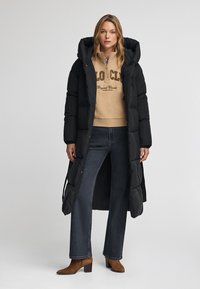 Mujer vestida con un abrigo de plumas largo y negro sobre una sudadera beige con cremallera, jeans oscuros y botines de tacón marrones, de pie contra un fondo sencillo.