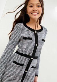 Sweter w różowym, czarnym i białym kolorze z teksturowanej tkaniny. Posiada okrągły dekolt, długie rękawy i czarne wykończenia z dekoracyjnymi guzikami.