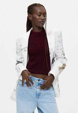Frau mit geflochtenem Haar, die ein burgunderfarbenes, strukturiertes Oberteil, einen weißen Blazer mit schwarzen Skizzen und hellblaue Jeans trägt, die Hände auf dem Jeansbund ruhend.