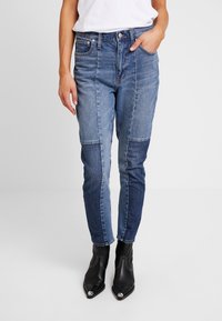 Jeans de mezclilla de parches en azul claro y oscuro, con un ajuste slim, que cuentan con bolsillos y cierre de botón, combinados con botas de tobillo puntiagudas negras.