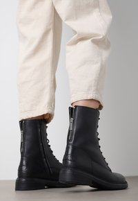Botas de tobillo de cuero negro con cierre de cordones y cremallera lateral, con una superficie texturizada y suelas de goma bajas. Llevadas con pantalones beige claros.