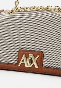 Béžová a hnědá kabelka s vzorem a zlatým řetízkovým popruhem, zdobená výrazným zlatým logem a hnědými koženými doplňky. Texturovaný povrch.