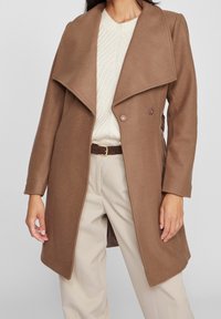 Brun stickad ullblandning med en stor krage, dubbelknäppt design och mjuk textur. Bärs över en krämfärgad kabelstickad tröja och beige byxor.