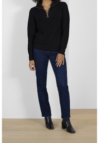 Maglione nero a coste con colletto alto e zip dorata, abbinato a jeans dritti blu scuro e stivali neri alla caviglia.