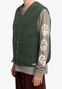 Groene gewatteerde vest met een gestructureerd oppervlak, knopen aan de voorkant en een label aan de onderkant. Draag je over een grijze langemouwen T-shirt met witte opdruk.