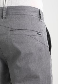 Pantalons gris en tissu lisse, dotés d'une poche arrière avec une étiquette noire Volcom. Le design comprend de subtils détails de couture.