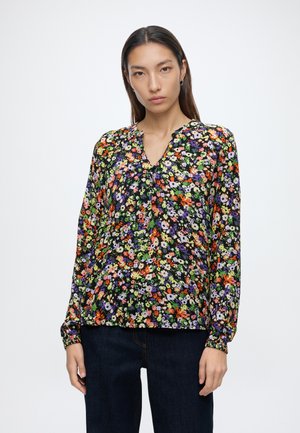 Jeune femme aux cheveux raides et foncés portant un chemisier à manches longues fleuri avec des fleurs orange, violettes, vertes et blanches, associé à un pantalon foncé.