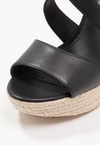 Sandalo in pelle nera con tacco, dotato di una larga fascia, punta aperta e suola in juta intrecciata con texture. Finitura liscia, design minimalista.