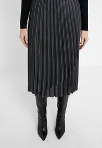 Falda midi negra con pliegues y brillo metálico, con rayas verticales y un dobladillo en ángulo. Combinada con botas altas de cuero negro con punta.