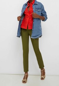 Vrouw in olijfgroene broek, rode blouse met knopen, spijkerjack en beige hakken met bandjes, poseert tegen een witte achtergrond.