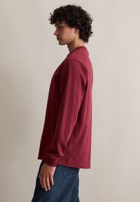 Langarm-T-Shirt aus burgunderfarbenem Baumwollstoff mit Rundhalsausschnitt, gerippten Bündchen und lockerer Passform. Kombiniert mit blauen Jeans.