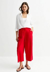 Pantalones culotte rojos, holgados y con cintura ajustable, combinados con una camisa blanca de botones y sandalias planas marrones. Textura de tela suave.