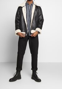Chaqueta de cuero negra con cuello y puños de piel sintética blanca, llevada sobre una camisa a rayas azul y blanca, combinada con pantalones negros remangados y botas.