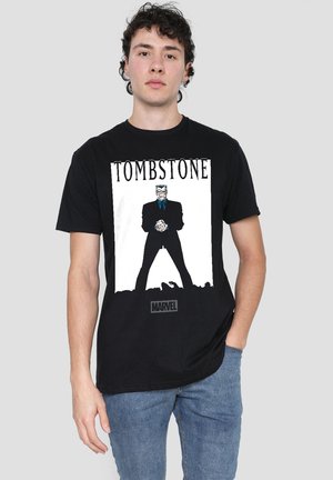 Young man wearing black Marvel Tombstone graphic t-shirt and blue jeans, standing against plain light gray background.