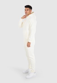 Conjunto de sudadera y pantalones jogger de color crema claro, hecho de una tela suave, con un bolsillo frontal y puños acanalados, combinado con zapatillas blancas.