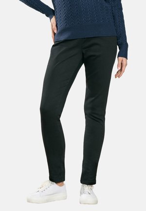 Person iført sorte slim-fit bukser, navy kabelstrikket sweater og hvide sneakers, står med den ene hånd på hoften.