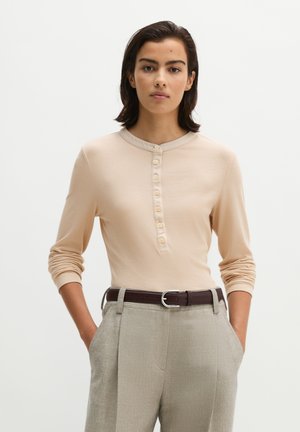 Vrouw met donker haar draagt een beige henley top met lange mouwen, ingestopt in lichtgrijze broek met hoge taille en een donkerbruine riem.