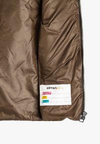 Interno di una giacca isolante marrone con texture liscia. Presenta una tasca etichetta con campi per nome e informazioni colorati. Dettaglio della cerniera nera.