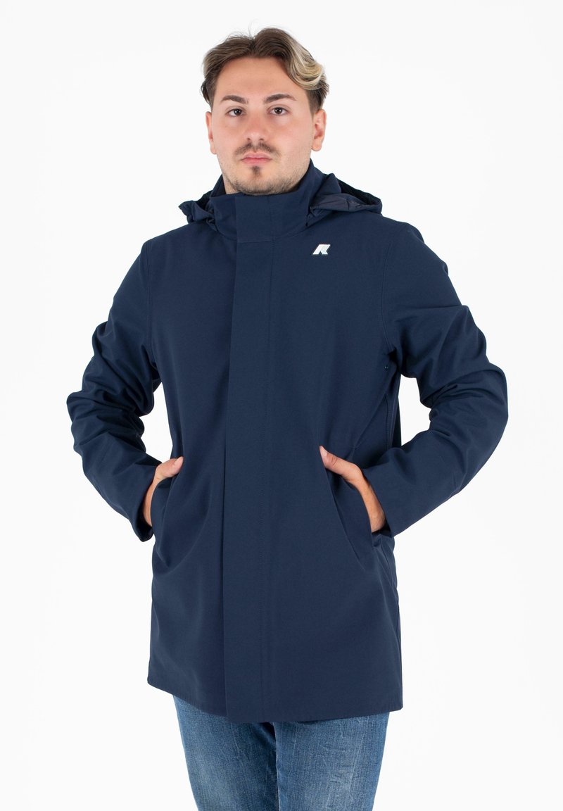 Giacca impermeabile blu navy con colletto alto, cappuccio regolabile, tasche laterali e logo sul petto. Texture del tessuto liscia.