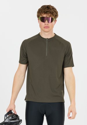 Nuori mies oliivinvärinen pyöräilypaita ja mustat shortsit, pitelee mustaa kypärää, pukeutuneena heijastaviin urheilulasit valkoista taustaa vasten.