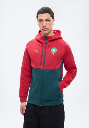 Junger Mann trägt einen rot-grünen Kapuzenpullover mit Reißverschluss, einem Puma-Logo und einem grünen Emblem auf der Brust, steht vor einem schlichten Hintergrund.
