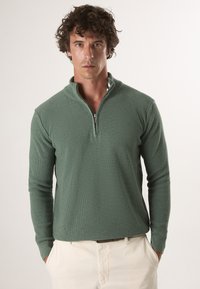 Grüner, gerippter Pullover mit einem Halbreissverschlusskragen und langen Ärmeln, aus strukturiertem Stoff gefertigt, kombiniert mit hellfarbigen Hosen.