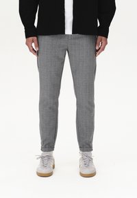 Man wearing gray tailored pants, white sneakers, white socks, and a black jacket over a white shirt standing on a white background.