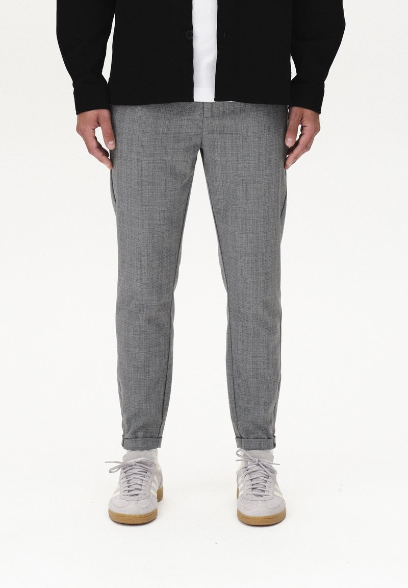 Man wearing gray tailored pants, white sneakers, white socks, and a black jacket over a white shirt standing on a white background.