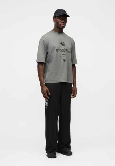 Sivá oversized tričko s grafikou "ULTRAS", kombinované s čiernymi voľnými nohavicami. Má krátke rukávy a okrúhly výstrih.