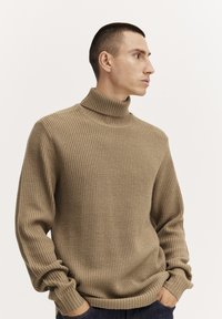 Beige ribbaava kauluspaita, jossa on teksturoitu neulos, rento malli ja tiukat hihansuut, päällä mallilla neutraalilla taustalla.