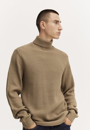 Beige bordázott garbó pulóver texturált kötésű, laza fazonú és illeszkedő mandzsettákkal, viselve egy modellen semleges háttér előtt.