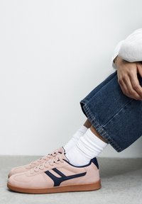 Rosa Nubuk-Sneaker mit marineblauen Akzenten, im klassischen Low-Top-Design, kombiniert mit weißen Crew-Socken und hochgekrempelten Jeans.