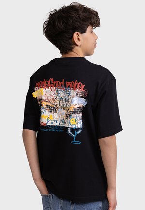 Dreng set fra siden iført sort T-shirt med farverigt graffitilignende tryk og tekst på ryggen, kombineret med blå jeans.