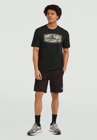 Young man standing wearing a black O'Neill t-shirt, black shorts, black socks, and gray sneakers against a plain light background.