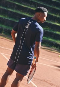 Ναυτικό αθλητικό μπλούζα με λευκό λογότυπο και λεπτομέρειες σε ρίγες, σε συνδυασμό με αντίστοιχα σορτς. Το άτομο κρατάει μία ρακέτα τένις σε μια χωμάτινη αρένα.