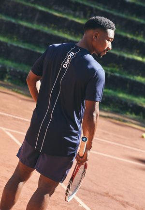 Marineblauwe sportshirt met wit logo en streepdetail, gecombineerd met bijpassende shorts. Persoon houdt een tennisracket vast op een gravelbaan.