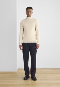Suéter de cuello alto beige en un diseño ajustado, combinado con pantalones oscuros de corte a medida y zapatos negros, contra un fondo minimalista.