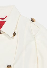 Λευκό παλτό τύπου trench με φαρδύ γιακά και καφέ κουμπιά σε σχέδιο χελώνας στο επιγονατίδιο του ώμου και στο εμπρόσθιο πάνελ.