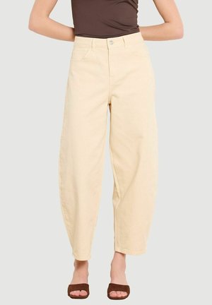 Naine, kes kannab kõrge vöökohaga, laiade säärega koorevärvseid pükse, üleni pruunist varrukatuta topist ja pruunidest libisemistest sandaalidest, seisab ühevärvilise tausta ees.